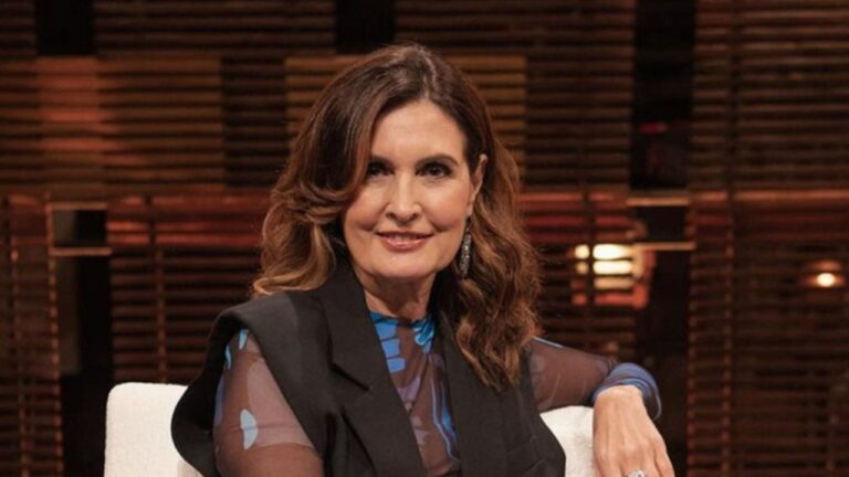 Fátima Bernardes sentada em estúdio com iluminação amena sorri levemente para a câmera, vestindo blusa estampada azul e colete preto, em um ambiente elegante com fundo de ripas de madeira