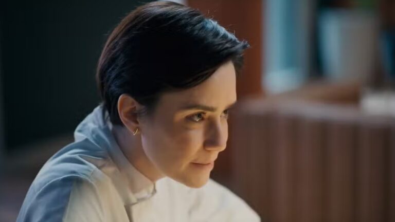 Fernanda Vasconcellos de cabelo curto e escuro veste camisa branca e observa algo com expressão concentrada em ambiente interno iluminado por luz natural