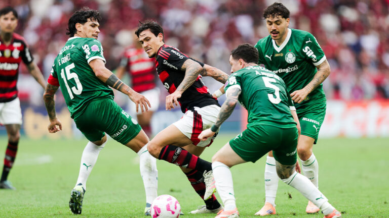 Quatro jogadores em campo, um do Flamengo com camisa preta e vermelha, e três do Palmeiras com camisa verde, disputando a bola