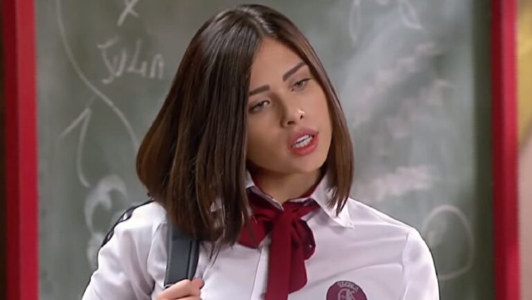 Jovem com uniforme escolar branco e gravata vinho aparece em sala de aula, com expressão séria e ligeiramente inclinada. Ao fundo, há um quadro com escritas e desenhos, sugerindo uma cena ambientada em um colégio