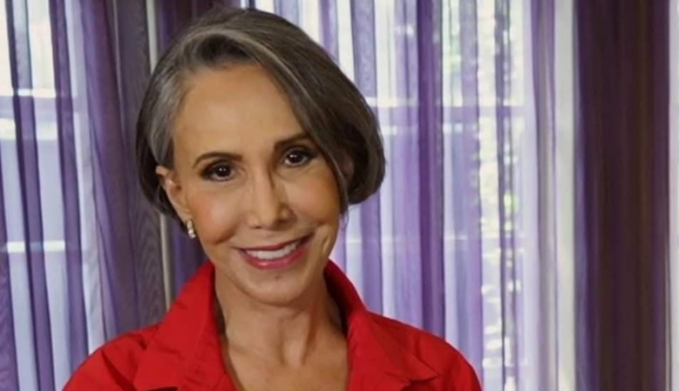 Mulher madura sorri em ambiente interno iluminado por luz natural. Ela tem cabelos curtos grisalhos penteados de lado e veste camisa vermelha. Ao fundo, cortinas lilás semi-transparentes deixam entrar a claridade