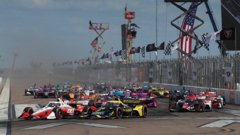 Carros alinham-se na largada de corrida da IndyCar com bandeiras quadriculadas ao fundo