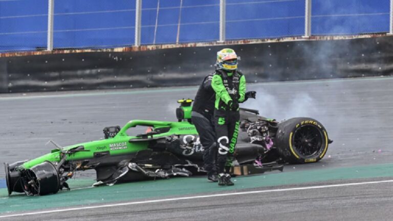A imagem mostra um carro de corrida totalmente destruído após um acidente na pista. O monoposto verde está partido ao meio, com rodas arrancadas e partes espalhadas, enquanto ainda sai fumaça da região traseira