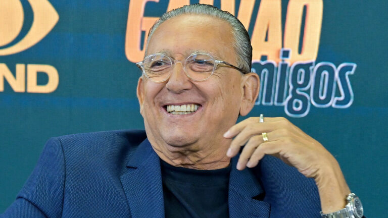 Narrador Galvão Bueno com blazer azul e óculos rindo em evento de lançamento do programa Galvão e Amigos na Band