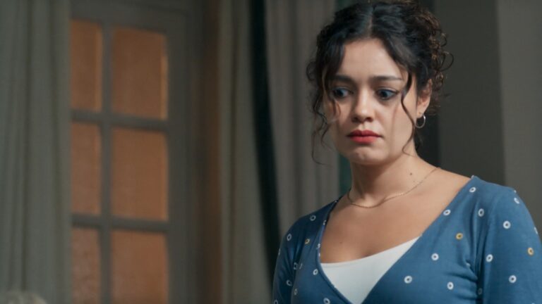 Gerluce, com expressão de preocupação está em ambiente interno com cortinas e janela ao fundo, vestindo blusa azul com pequenos círculos brancos e amarelos sobre regata branca em cena da novela Três Graças; personagem é vivida pela atriz Sophie Charlotte