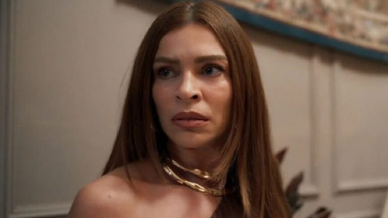 A imagem mostra uma mulher de expressão séria, com cabelo liso e castanho, usando um colar dourado espesso e vestido sem alças. Ela está em um ambiente interno, com uma parede clara e parte de uma tapeçaria visível ao fundo