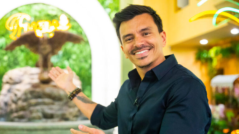 Autor Gustavo Reiz usando camisa preta e pulseiras, gesticulando com a mão direita em frente a uma fonte decorativa