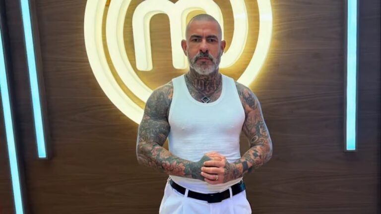 Homem tatuado de regata branca posa diante do símbolo iluminado do MasterChef em cenário de estúdio