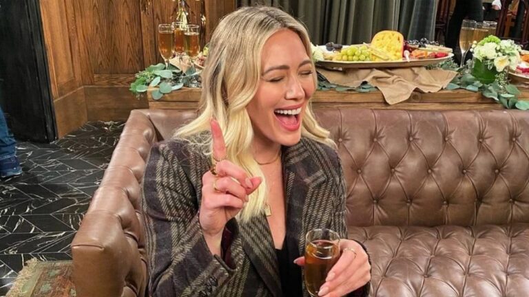 A imagem mostra uma mulher sorridente sentada em um sofá de couro marrom, vestindo um blazer xadrez. Ela segura um copo de bebida em uma mão e levanta o dedo indicador da outra, em um gesto animado. Ao fundo, há uma mesa com taças, frutas e queijos, sugerindo um ambiente de confraternização ou celebração