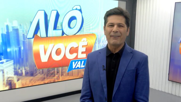 A imagem mostra um apresentador de televisão em um estúdio, vestindo um paletó azul e camisa preta. Ao fundo, há uma tela grande com o logotipo do programa Alô Você Vale, que possui letras em azul e laranja sobre uma imagem de cidade desfocada. O cenário é moderno e bem iluminado, típico de programas de variedades ou jornalísticos