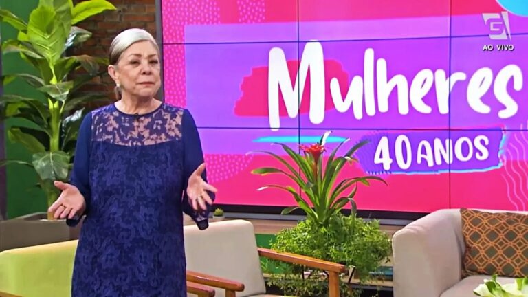 A imagem mostra uma mulher de cabelos presos e roupa azul rendada apresentando um programa de TV. Ela está em um estúdio colorido, com o logo “Mulheres 40 anos” exibido em um grande painel ao fundo. O cenário tem plantas e poltronas, transmitindo um ambiente acolhedor e descontraído típico de programas de variedades