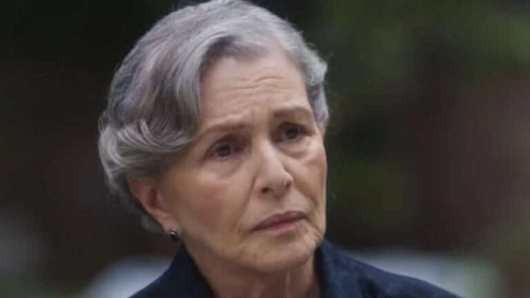 Mulher idosa com cabelos grisalhos curtos e expressão séria aparece em cena externa, sob luz natural e com fundo desfocado