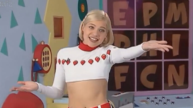 Jackeline Petkovic no Bom dia & Cia com top branco e mini saia vermelha, gesticulando no cenário com o painel de letras