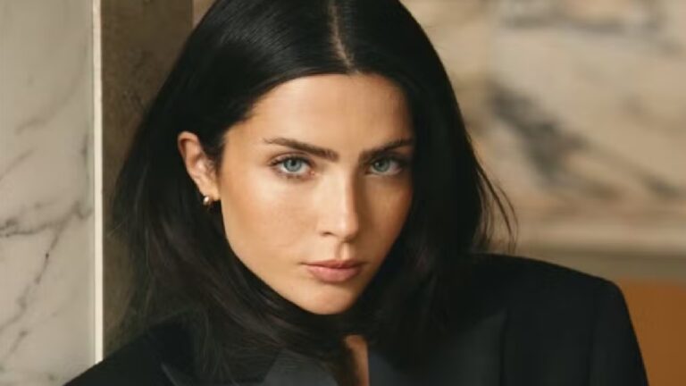 A imagem mostra uma mulher de cabelos escuros e lisos, pele clara e olhos claros, olhando diretamente para a câmera com uma expressão séria. Ela está usando um blazer preto, e o fundo apresenta um tom neutro com detalhes em mármore, criando um ambiente elegante e sofisticado