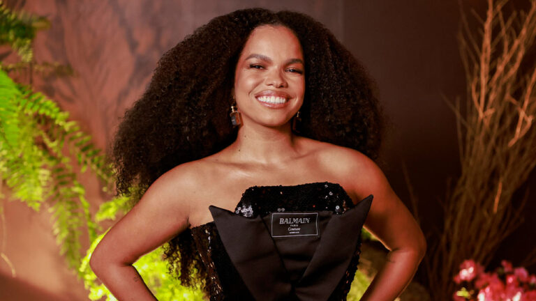 Atriz Jeniffer Nascimento sorrindo com cabelo cacheado volumoso, usando vestido preto de lantejoulas e laço frontal