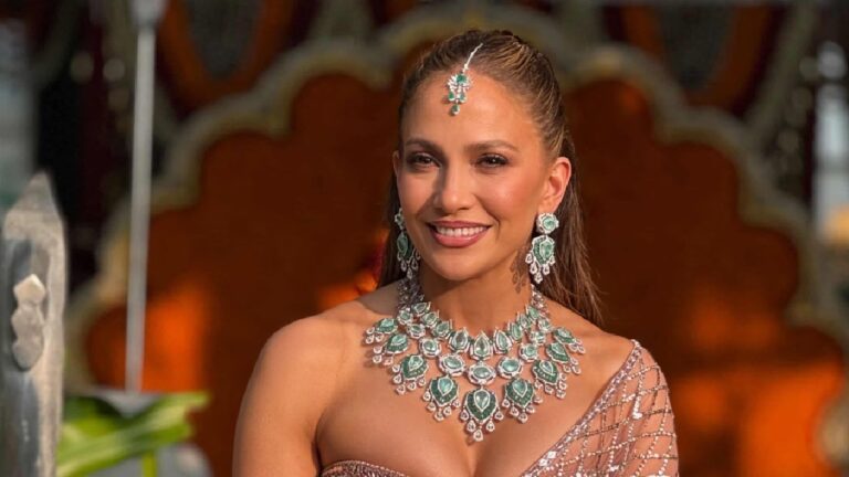 Mulher sorrindo com colar e brincos de pedras verdes em ambiente decorado com painel alaranjado ao fundo