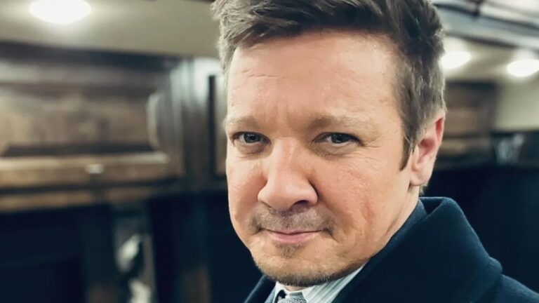 Homem de expressão séria posa para a câmera em ambiente interno, vestindo paletó escuro e camisa com gravata, sob iluminação suave