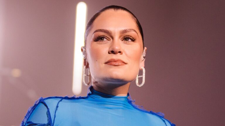 A imagem mostra uma mulher em um palco ou ambiente de apresentação, usando um vestido azul vibrante e brincos grandes e brilhantes em formato retangular. Ela está com o cabelo preso em um coque bem alinhado, usando maquiagem suave porém elegante, com foco nos olhos e lábios gloss. Também é possível ver um ponto de luz ao fundo e um pequeno fone de retorno no ouvido, típico de performances musicais