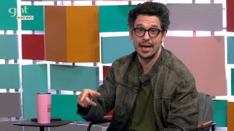 Homem de óculos e jaqueta verde fala durante programa de TV ao vivo no canal GNT, com cenário colorido ao fundo