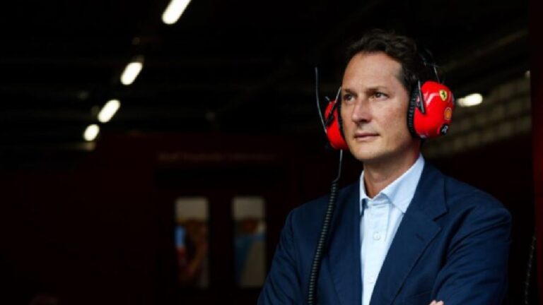 Homem com expressão concentrada usa protetores auriculares vermelhos com logotipos automotivos e terno azul-escuro, observando algo à distância em ambiente de boxes de corrida iluminado por luzes artificiais