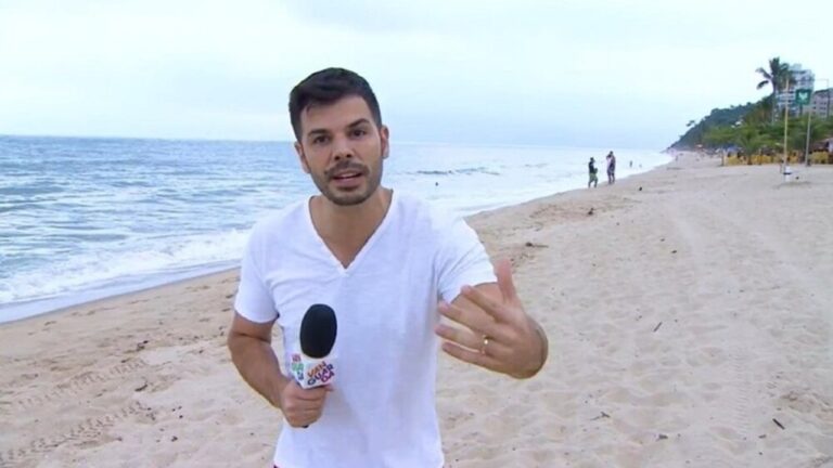 Jonas Almeida segura microfone e fala diante da câmera em uma praia, com o mar e algumas pessoas ao fundo, em dia nublado