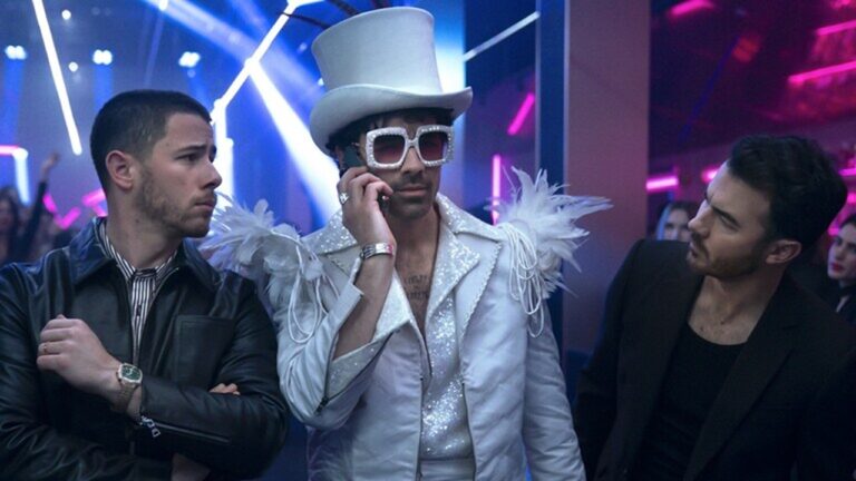 Três homens caminham juntos em uma balada iluminada por luzes de néon. O homem ao centro veste uma fantasia chamativa totalmente branca, com casaco brilhante, ombreiras de plumas, cartola alta e óculos quadrados com brilho, enquanto fala ao celular. Os dois homens ao lado usam roupas escuras e o observam com expressões de surpresa e curiosidade. O ambiente ao fundo está cheio de pessoas e luzes vibrantes, criando um clima de festa