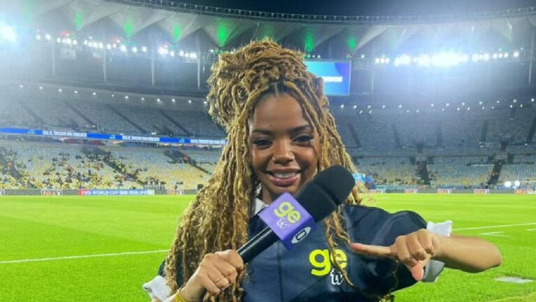 A imagem mostra uma repórter esportiva sorridente em um estádio de futebol. Ela segura um microfone com o logotipo do ge.globo e usa uma jaqueta da emissora. O campo verde e as arquibancadas iluminadas ao fundo indicam que ela está cobrindo um evento esportivo, possivelmente uma partida noturna