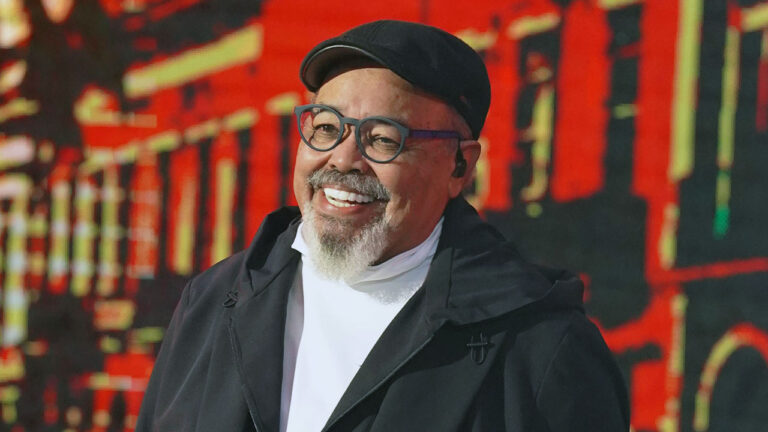 Cantor Jorge Aragão sorridente, com cavanhaque branco, óculos de aro escuro e boné preto, em show em palco com LED vermelho