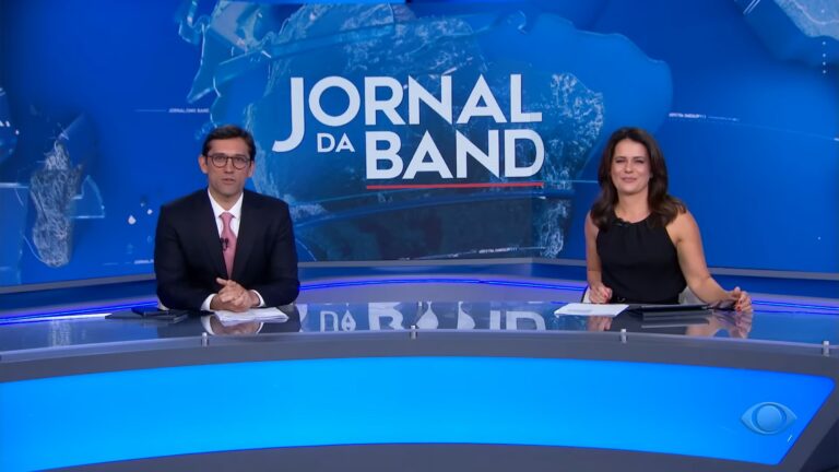 Dois jornalistas sentados atrás da bancada do Jornal da Band durante a apresentação do telejornal, com o logotipo do programa em destaque em um grande painel azul ao fundo e o estúdio iluminado em tons de azul e cinza