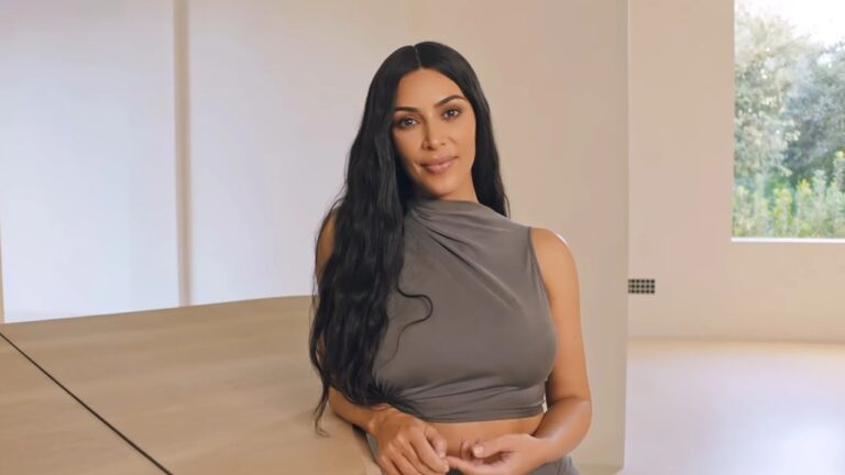 A imagem mostra uma mulher de cabelos longos e pretos, posicionada em um ambiente interno amplo e minimalista, com paredes claras e janelas grandes ao fundo. Ela está apoiada em uma superfície de madeira clara e veste um top cinza de modelagem elegante. A expressão é serena e simpática, com postura relaxada em um cenário bem iluminado por luz natural.