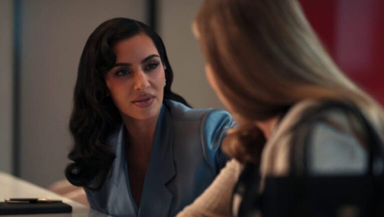 A imagem mostra duas mulheres em uma conversa em ambiente interno. A mulher à esquerda veste um blazer azul e tem cabelo escuro e ondulado, enquanto a outra, de costas, tem cabelo loiro e liso. A cena tem uma iluminação suave, indicando que pode ser parte de uma produção cinematográfica ou série