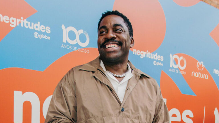 Lázaro Ramos sorrindo de bigode, vestindo jaqueta bege sobre camisa clara em evento Negritudes da Globo