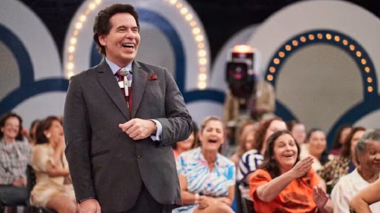 Leandro Hassum de terno escuro sorri amplamente enquanto apresenta um programa de auditório, com público rindo e aplaudindo ao fundo em cenário colorido e iluminado