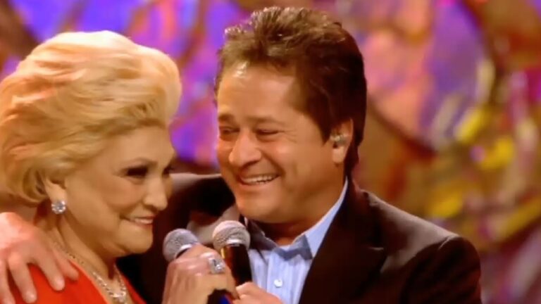 A imagem mostra um homem e uma mulher sorridentes se abraçando enquanto cantam em um palco colorido. Ambos seguram microfones e aparentam estar emocionados durante a apresentação. A mulher, de cabelos loiros e penteado volumoso, usa roupa vermelha elegante e joias brilhantes