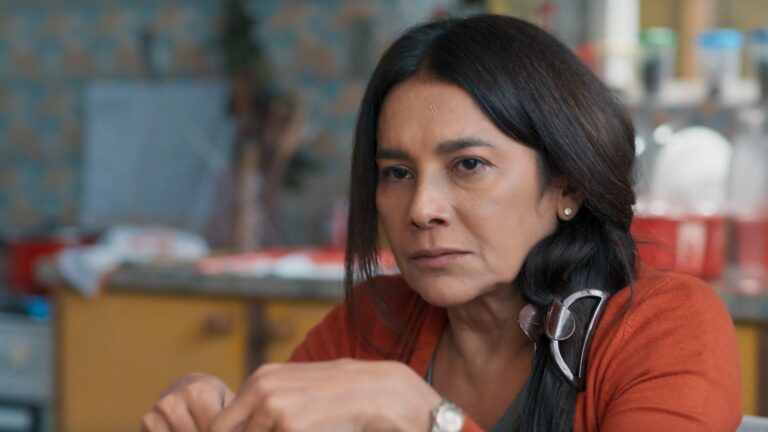 Dira Paes, com expressão séria e olhar concentrado, aparece sentada em ambiente doméstico com utensílios de cozinha ao fundo, usando cardigã laranja e prendedor grande no cabelo em cena da novela Três Graças, onde interpreta a personagem Lígia