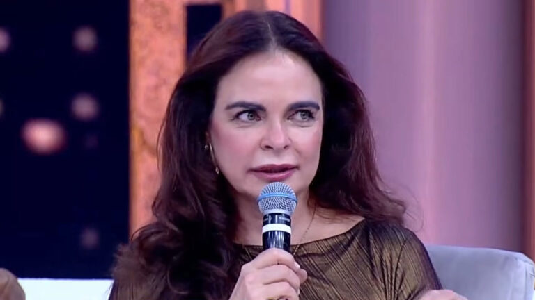 Liliane Ventura com cabelo castanho longo, usando blusa dourada e falando ao microfone em participação em programa de TV