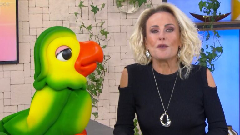 A imagem mostra Ana Maria Braga, uma apresentadora de TV loira, usando um vestido preto com ombros à mostra e colar prateado, ao lado de Louro José, um boneco colorido de papagaio com bico laranja e corpo verde e amarelo. Eles estão em um estúdio decorado com plantas e parede de pedra clara, no cenário do programa matinal Mais Você, da TV Globo