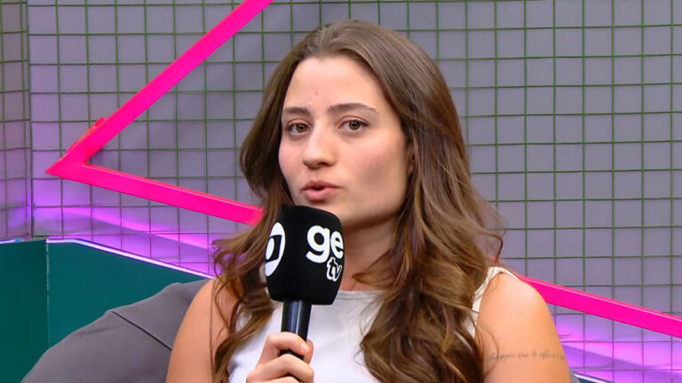 Luana Maluf falando e segurando microfone GE TV, com cabelo castanho ondulado, em estúdio de cores vibrantes