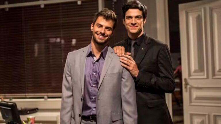 Dois homens posam juntos em um ambiente interno elegante, parecendo um escritório. O homem à frente usa um terno cinza claro com camisa roxa e sorri para a câmera. Atrás dele, outro homem veste um terno preto e apoia as mãos nos ombros do primeiro, também sorrindo. Ao fundo, há persianas escuras e uma porta branca parcialmente aberta, criando um clima formal e acolhedor