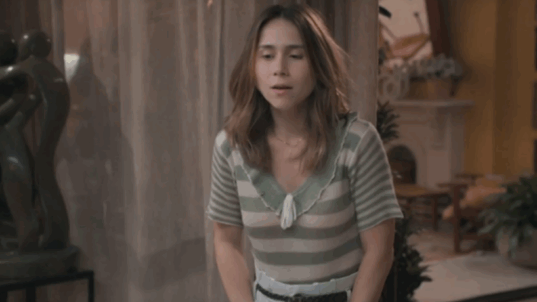 Mulher com expressão de surpresa ou inquietação está em uma sala decorada, vestindo blusa listrada em tons claros. Ao fundo, há cortinas translúcidas, plantas e uma escultura escura, compondo um ambiente acolhedor e elegante