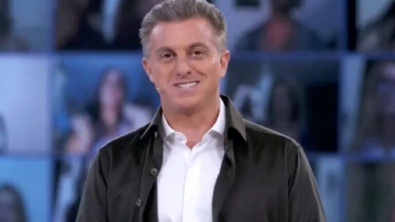 A imagem mostra um homem sorridente, usando camisa branca e jaqueta escura, em um estúdio de televisão com fundo azul e imagens desfocadas de pessoas em telas. Ele usa microfone de lapela e parece estar apresentando um programa ao vivo