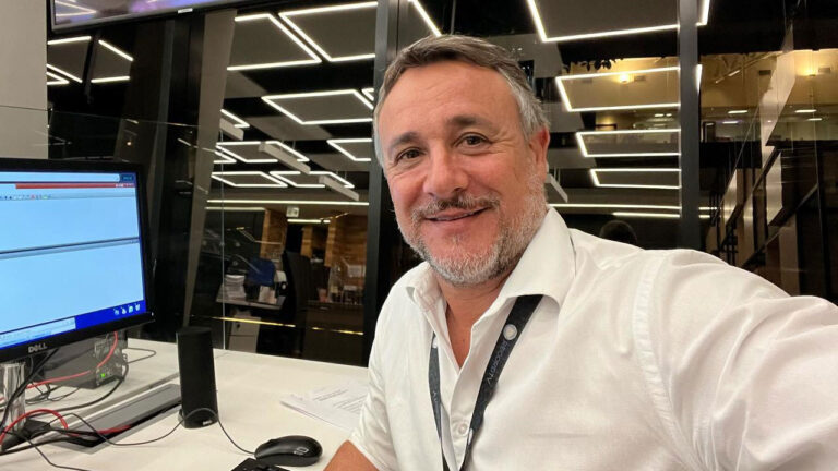 Lucio Sturm sorrindo em estação de trabalho na redação da Record, vestindo camisa social branca, com computador à esquerda