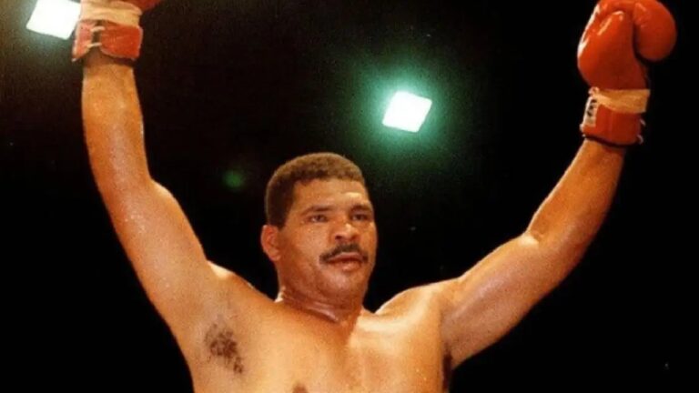 Boxeador de luvas vermelhas ergue os braços em sinal de vitória, suado e determinado, sob luzes intensas de um ringue de luta