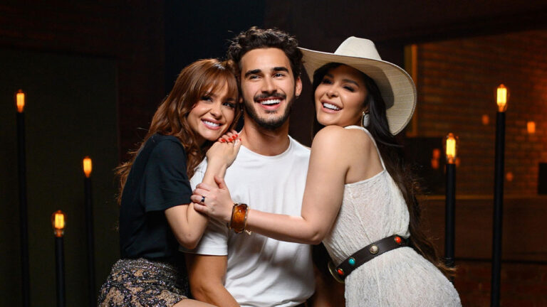 Maiara e Maraisa posando sorridentes ao lado de João Raul interpretado por Filipe Bragança em ambiente com iluminação quente e decorado com lâmpadas de filamento