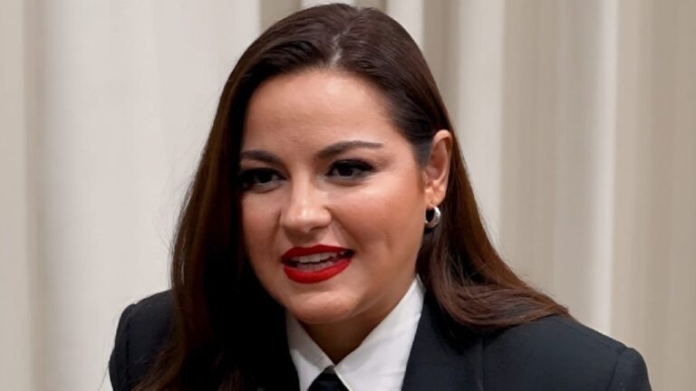 A imagem mostra uma mulher de cabelos castanhos lisos e maquiagem marcante, com batom vermelho e delineador preto. Ela está vestindo um terno preto com camisa branca e gravata, transmitindo um visual elegante e formal. O fundo é neutro, com cortinas claras