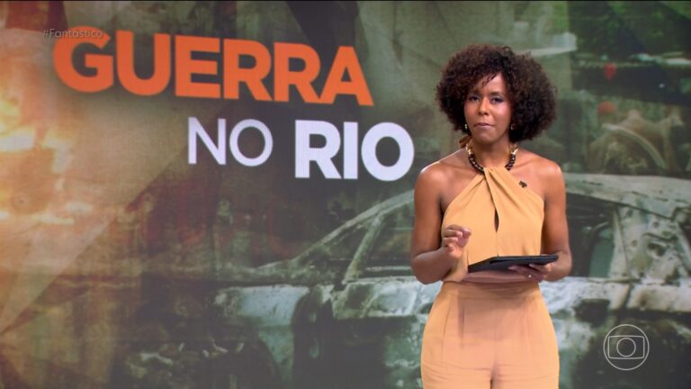 Maju Coutinho apresenta o programa Fantástico em estúdio da TV Globo, vestindo macacão bege e colar escuro, segurando um tablet enquanto aparece diante de um telão com a manchete “Guerra no Rio” e imagem de carro destruído ao fundo