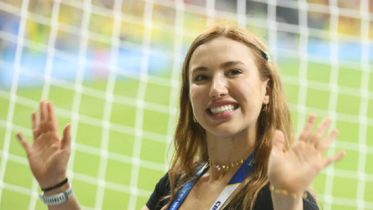 Mulher sorri e acena em frente a uma rede de gol em campo de futebol, usando crachá no pescoço e pulseiras nos braços. A cena transmite alegria em um ambiente esportivo, possivelmente durante um evento ou cobertura de jogo