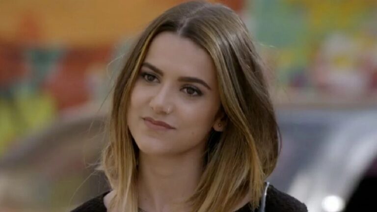 Mulher com cabelo castanho e mechas loiras olha ligeiramente para o lado com expressão pensativa, em cena externa com fundo colorido e desfocado