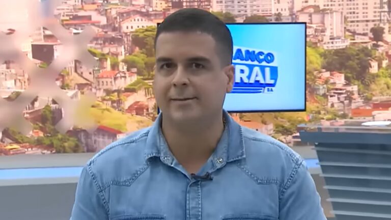 A imagem mostra o apresentador Marcelo Castro, do jornal Alô Juca, usando uma camisa jeans clara e microfone preso à gola. Ao fundo, há um painel com imagem panorâmica de uma cidade e uma tela exibindo o logotipo do programa Balanço Geral BA, em reportagem do Domingo Espetacular. O ambiente é bem iluminado e moderno, característico de um noticiário televisivo