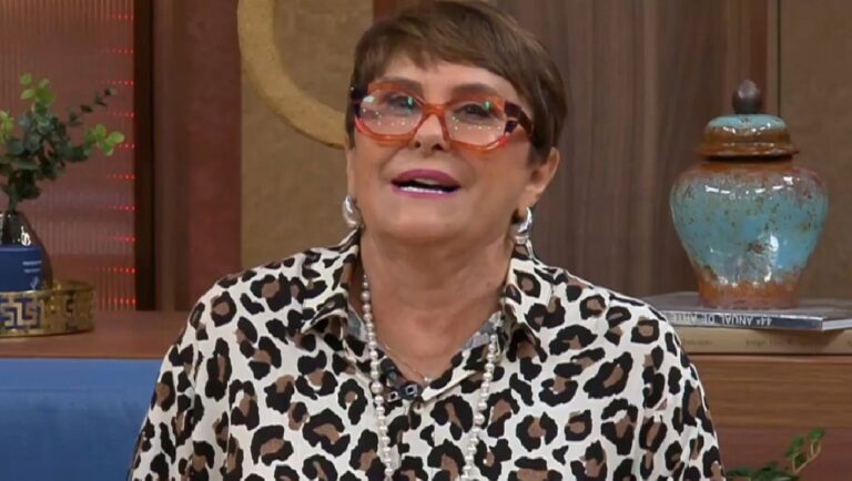 Mulher madura usando camisa estampada de oncinha, colar de pérolas e óculos de armação grossa em tons alaranjados aparece sorrindo em um estúdio decorado com plantas, livros e objetos de cerâmica ao fundo